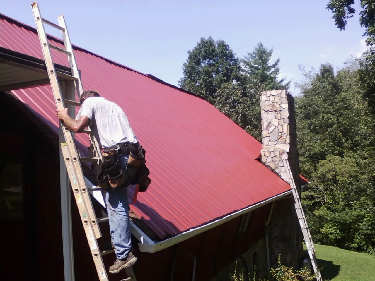 Roofer on a ladder beside a red metal roof and stone chimney — Soffit & Fascia Repair by Roofing Brothers in Kelso